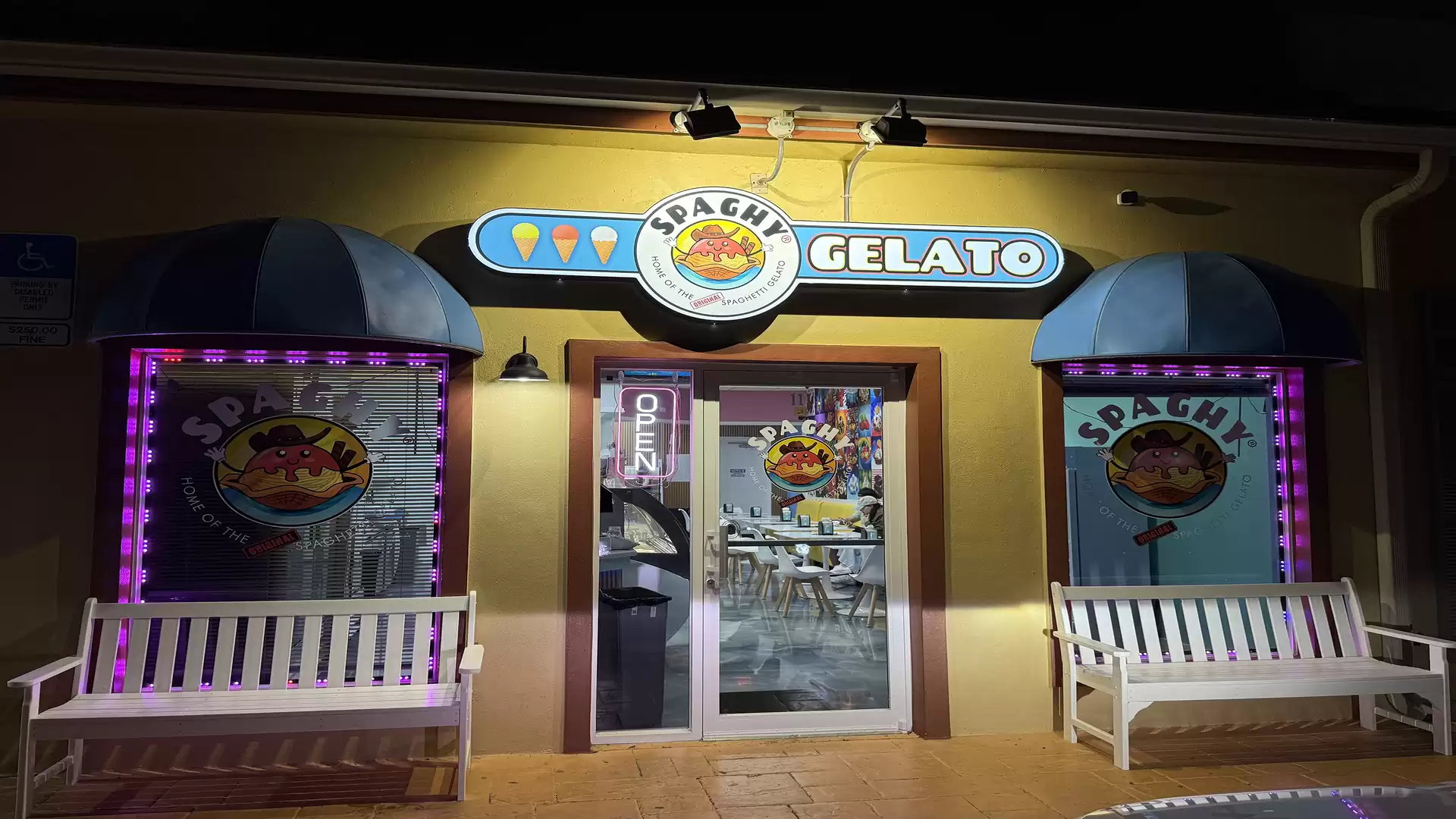 Image of an illuminated ice cream shop named 'Spaghetty Gelato', painted windows and benches in front of the entrance.