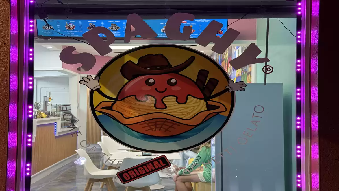 A lit storefront featuring a colorful logo of a smiling cartoon character in a cowboy hat sitting on a pile of spaghetti. The word 'Spaghy' is displayed above.