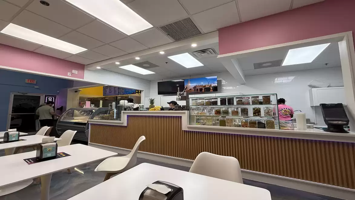 Interior view of a modern café with white seating and bright ceiling lights. The counter is equipped with glass displays showcasing various dishes.