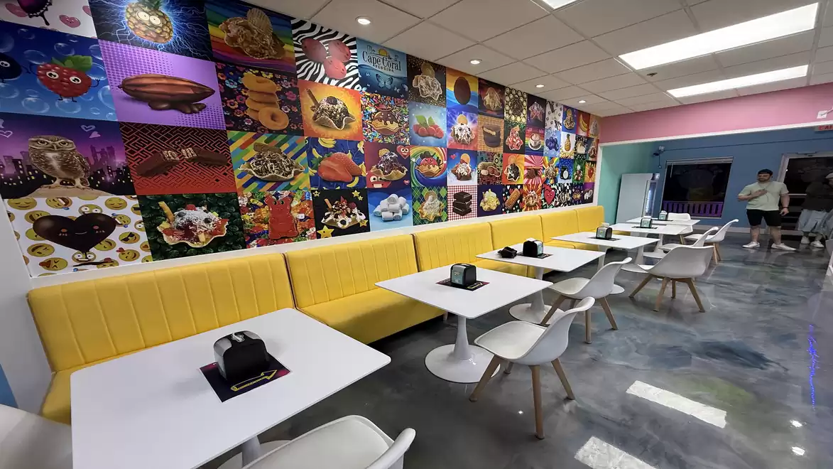 A modern café with a yellow bench, white tables, and chairs. The colorful wall is covered with various vibrant artworks.
