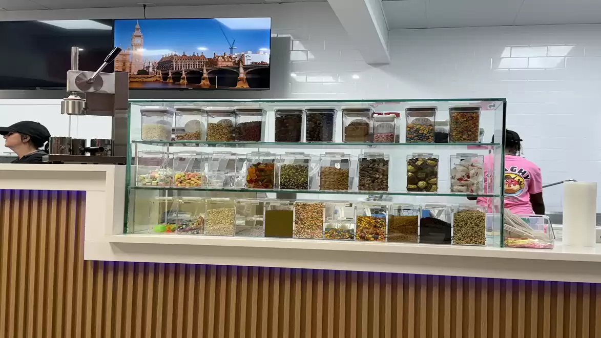 Interior of a modern tea shop with tea pots and containers on glass shelves, TV in the background displaying a cityscape.