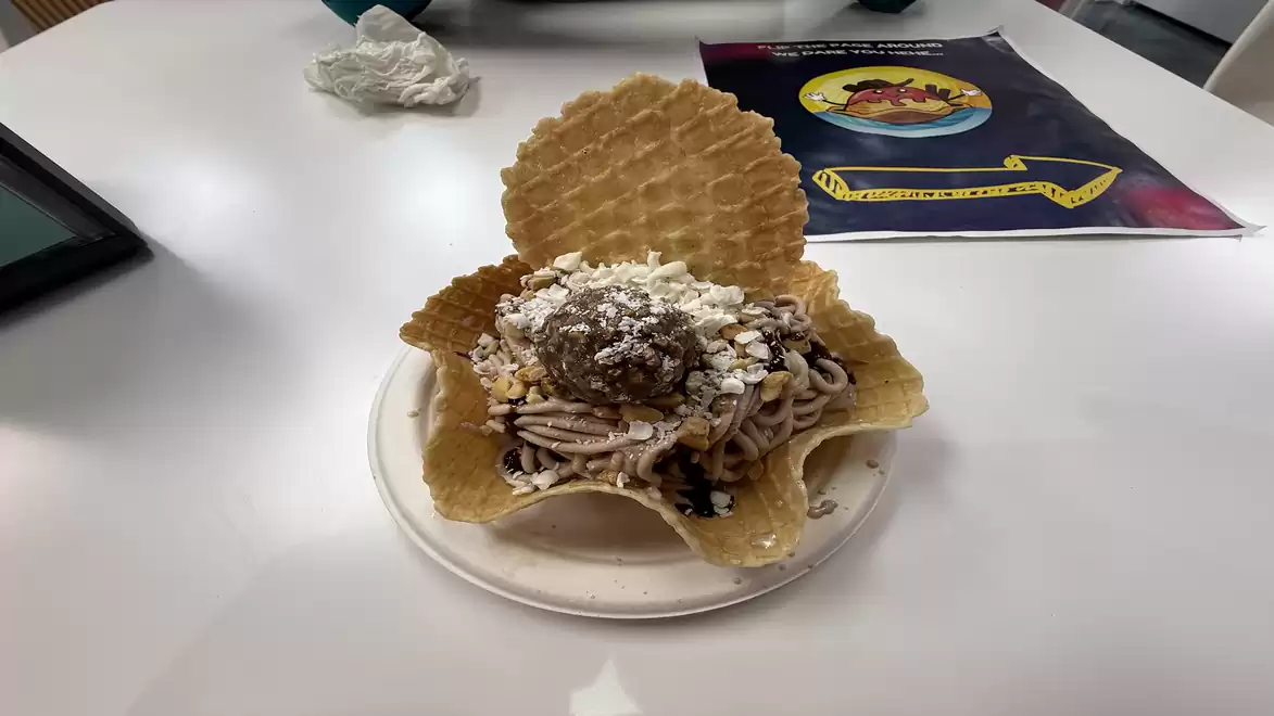 A plate of spaghetti ice cream served in a waffle bowl on a white table. In the background, a folded paper towel and a colorful poster are visible.