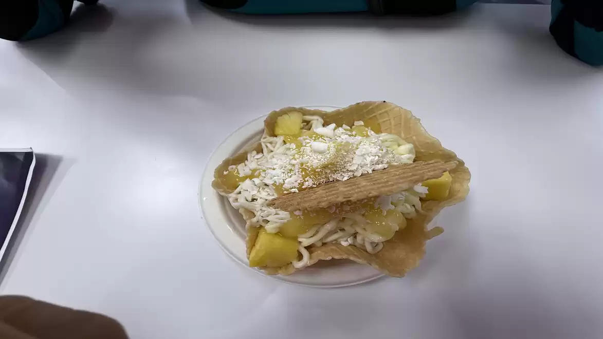 A plate of spaghetti ice cream topped with pineapple pieces, a waffle, and grated white chocolate, on a white table.