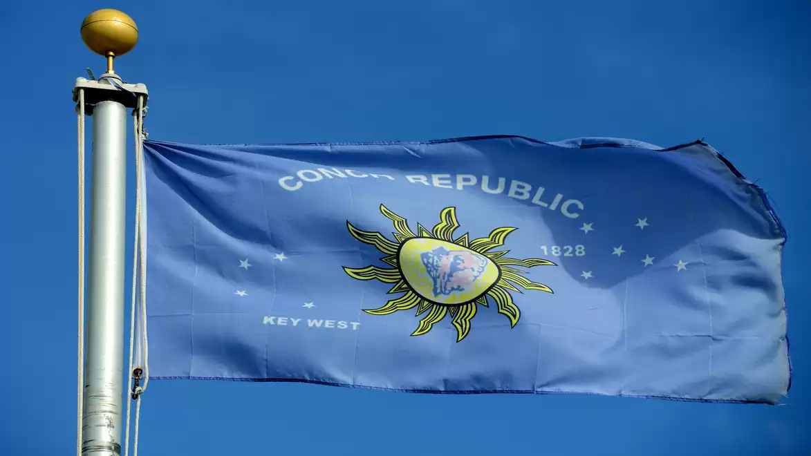 Blue and white Conch Republic flag with sun logo, waving in the wind against a clear sky.
