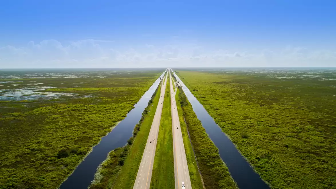 Aerial view of the Alligator Alley, a long, straight road surrounded by green landscape and two parallel water channels under a blue sky.