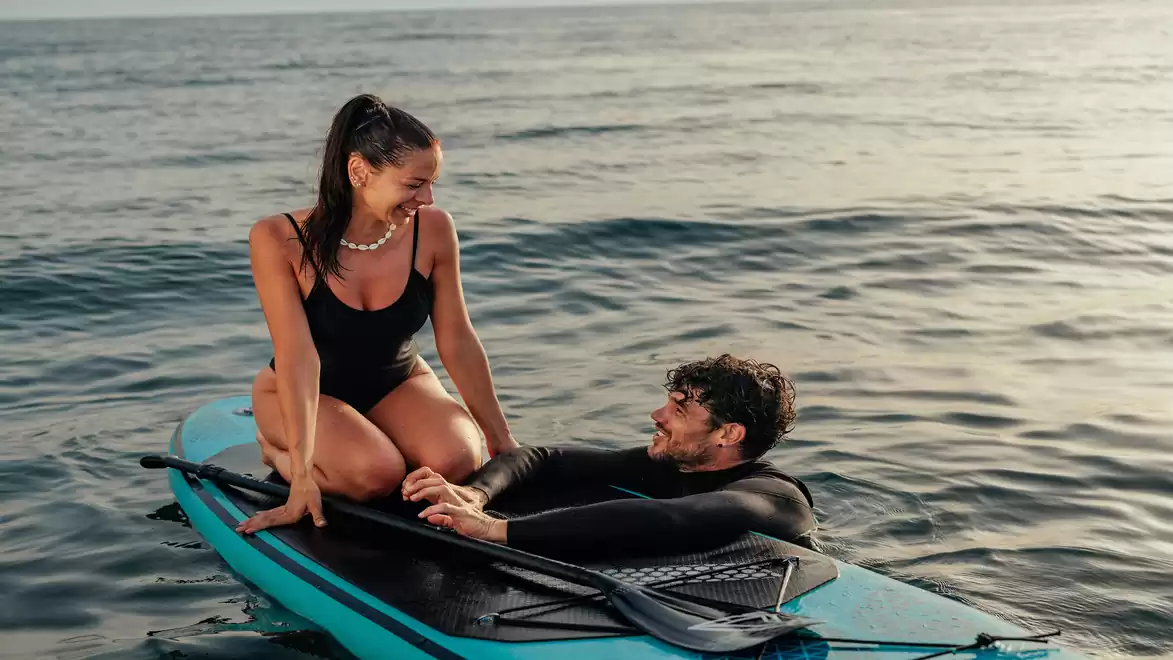 A man and a woman enjoying a relaxed time on a surfboard in the sea. The woman sits on the board while the man leans in the water.