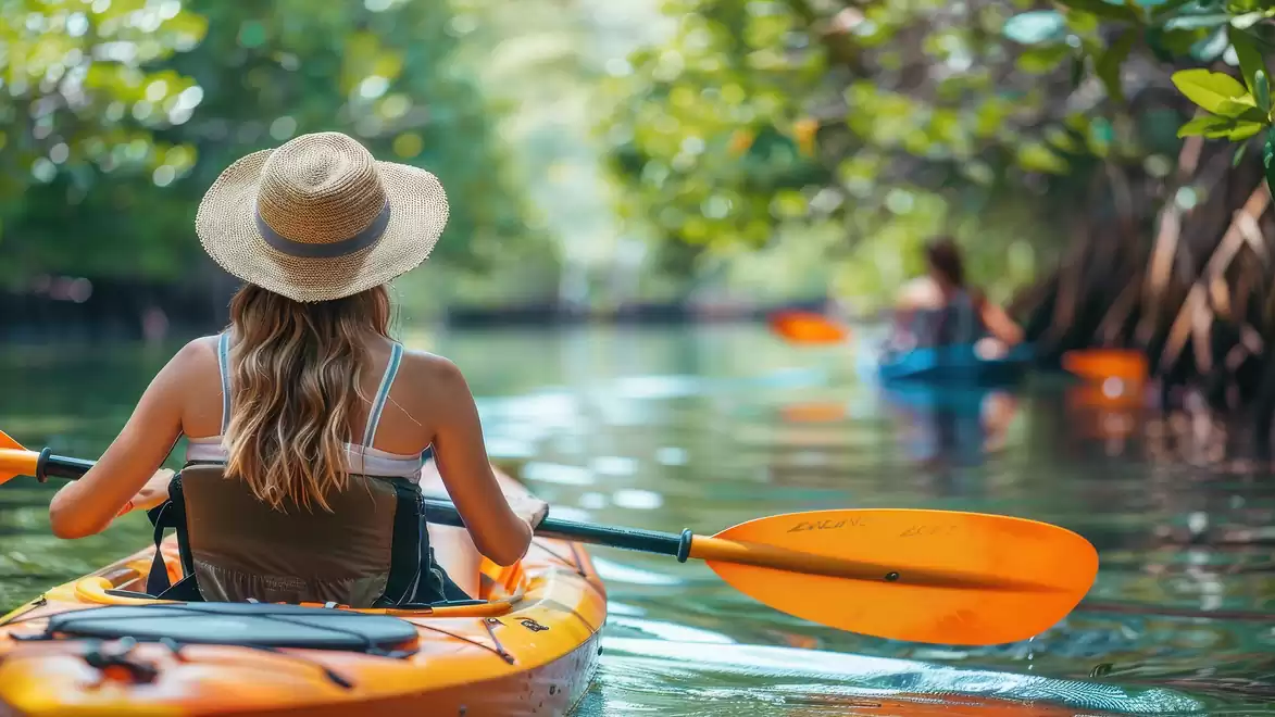 Woman with a straw hat paddling an orange kayak on a river through dense green vegetation.