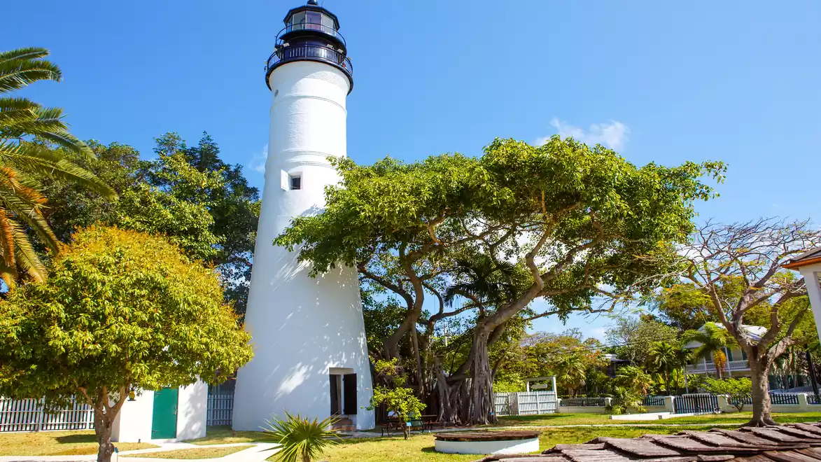 A white lighthouse stands in a sunny park with green trees and blue sky in Key West.