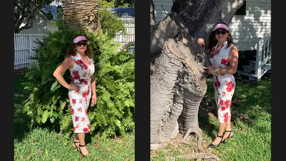 Woman in a white dress with red floral pattern and sunglasses posing next to a tree outdoors.