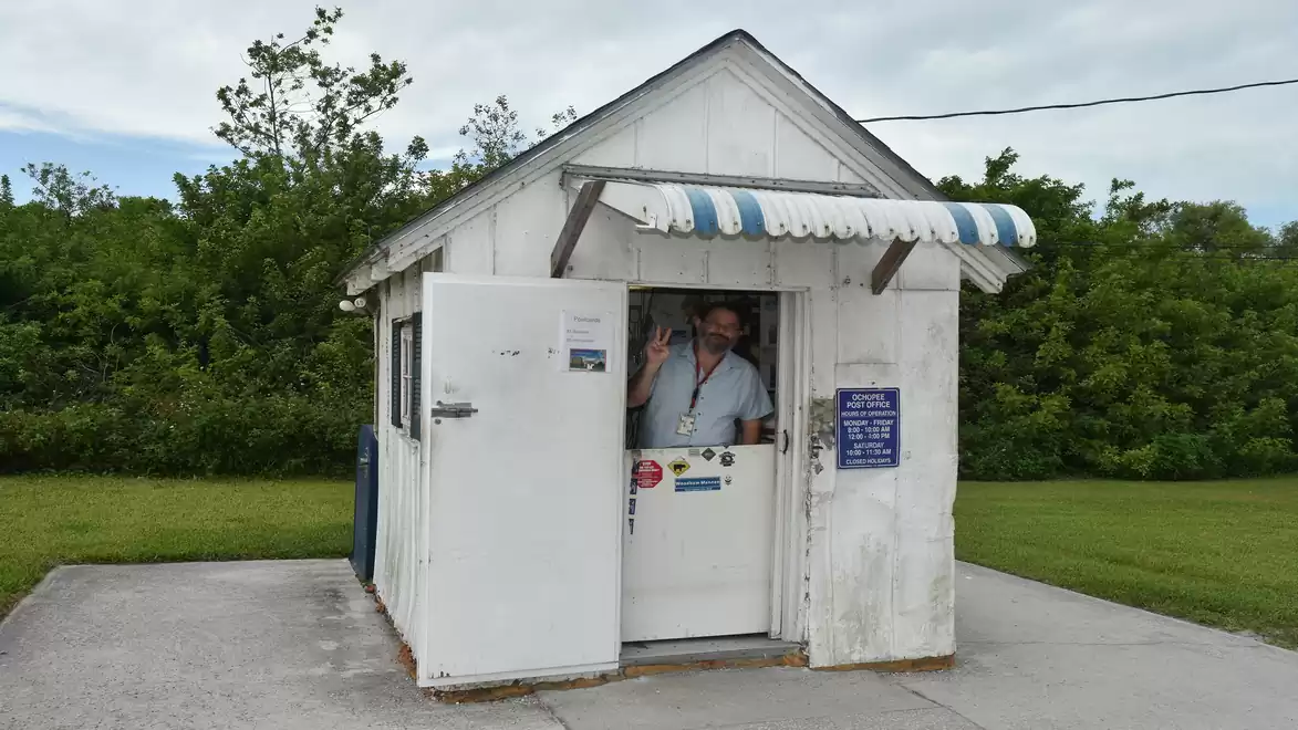 A small white building with a blue and white awning. A person is standing in the open door, waving. Green vegetation is visible in the background.