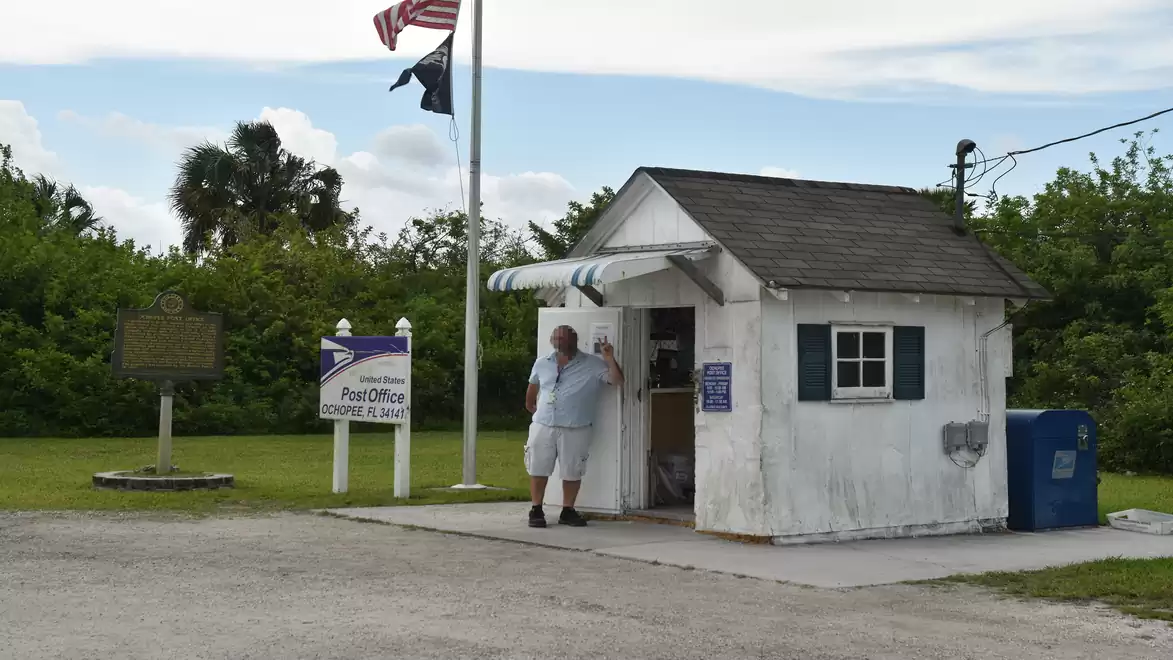 Small white post office with a shingle roof, situated in a green environment. A person stands in front of the building, the sky is slightly cloudy.