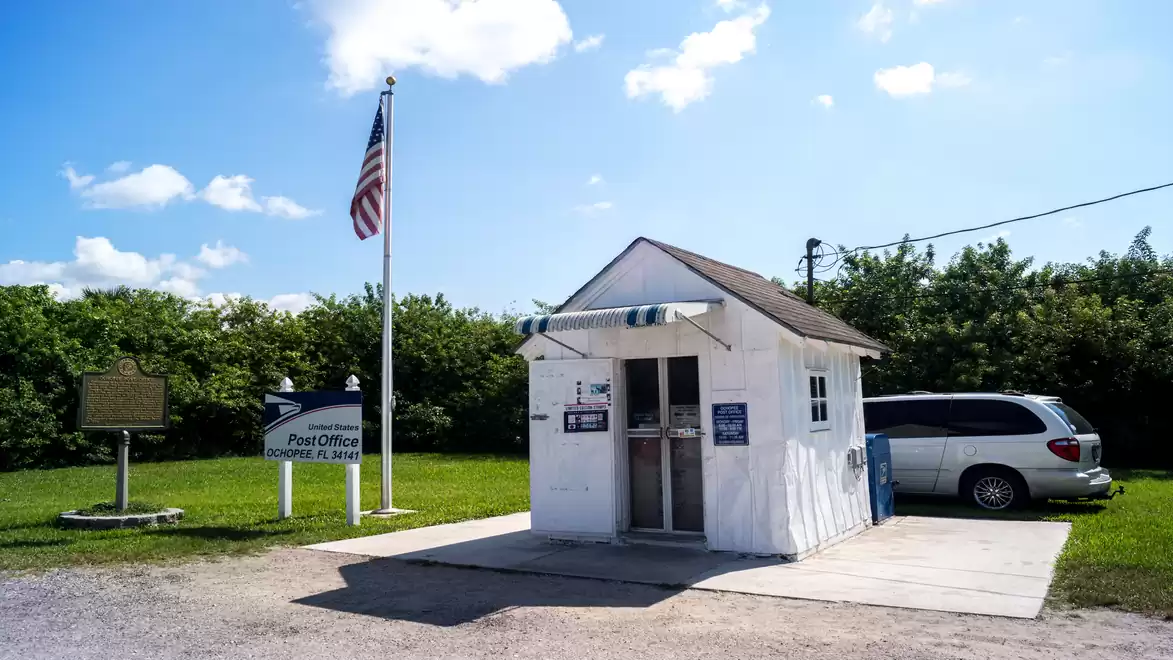 Small white post office with American flag and parked car on a sunny day.