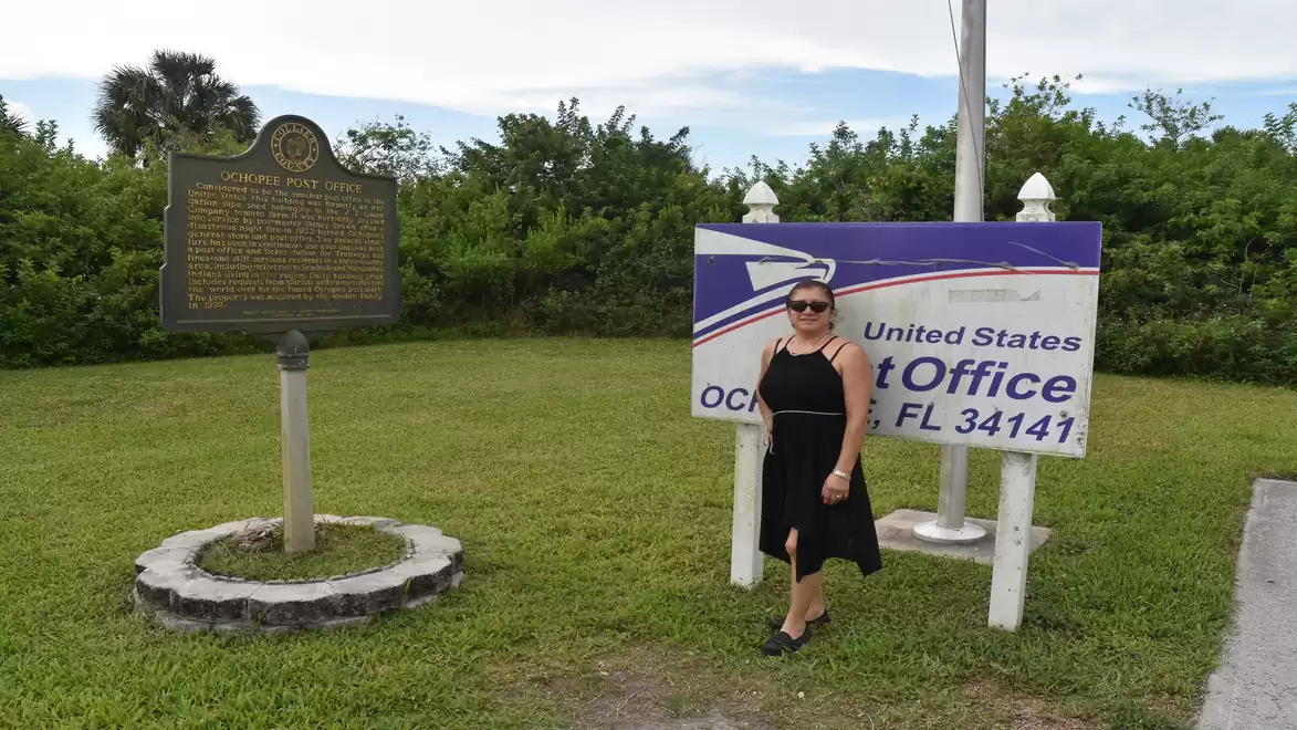 Woman in black dress standing next to a historical marker and a US Post Office sign in Ochopee, Florida.