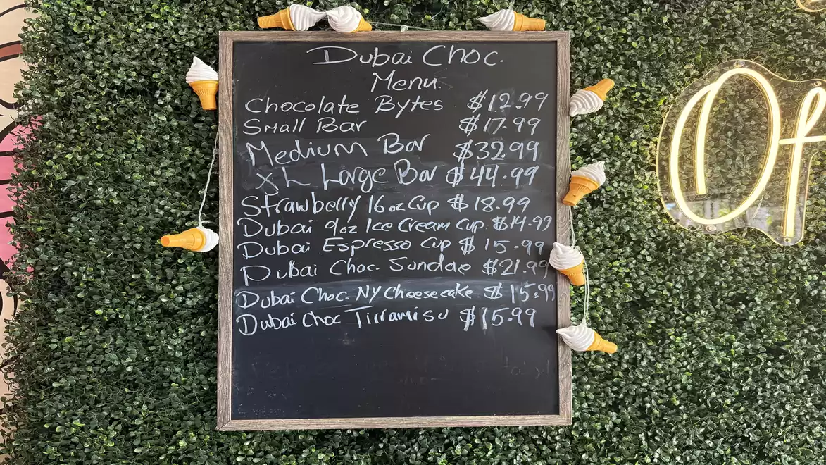 A menu board featuring Dubai Choc's items, surrounded by artistic green foliage and small orange and white decorative ice cream cones. The prices are listed in dollars, featuring various chocolate products.