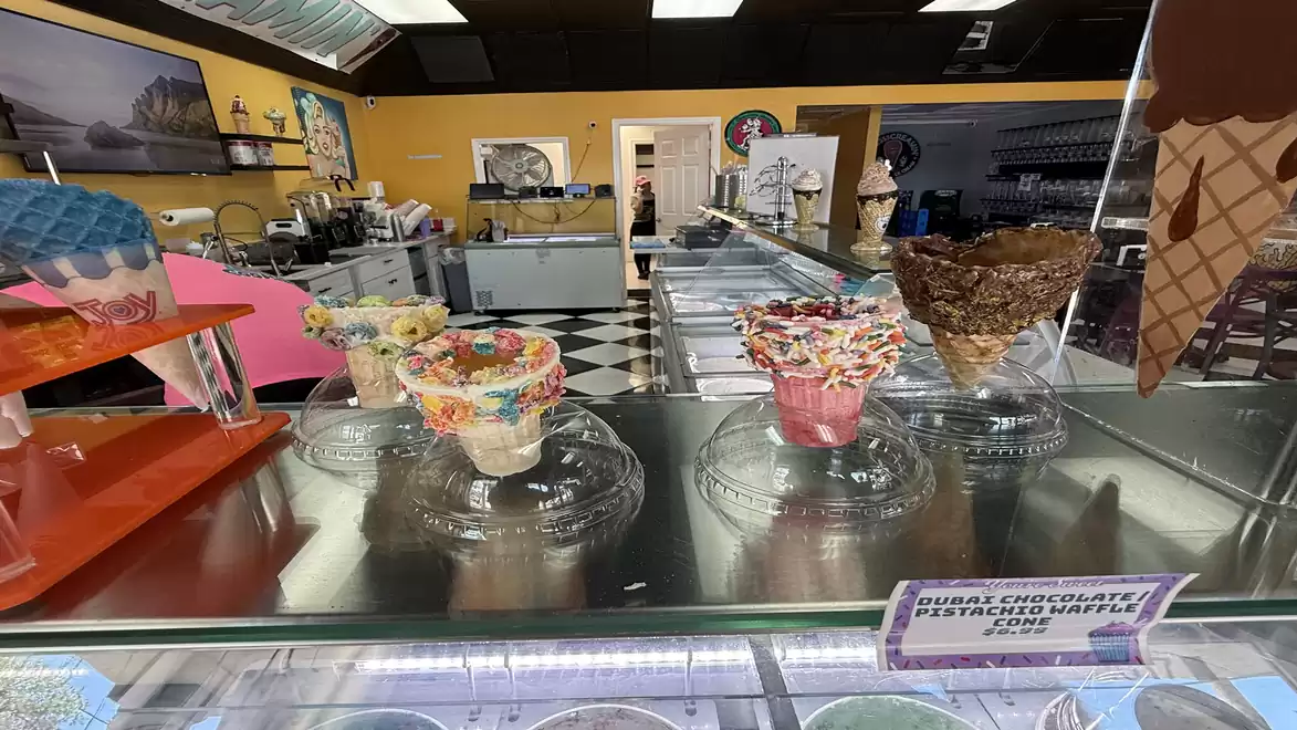 An ice cream parlor with various colorful and decorated ice cream cones on a counter. Ice cream machines and colorful walls can be seen in the background.