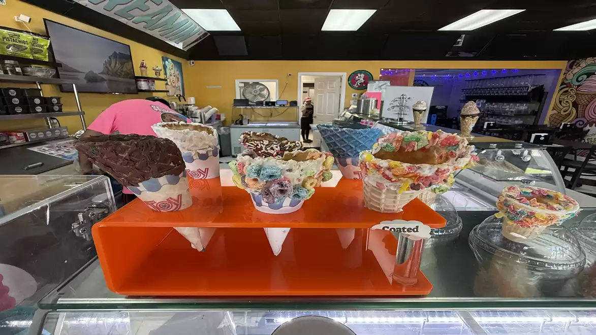 An ice cream shop featuring a selection of decorated cones displayed on an orange stand. Shelves with ice cream ingredients and decorations are visible in the background.