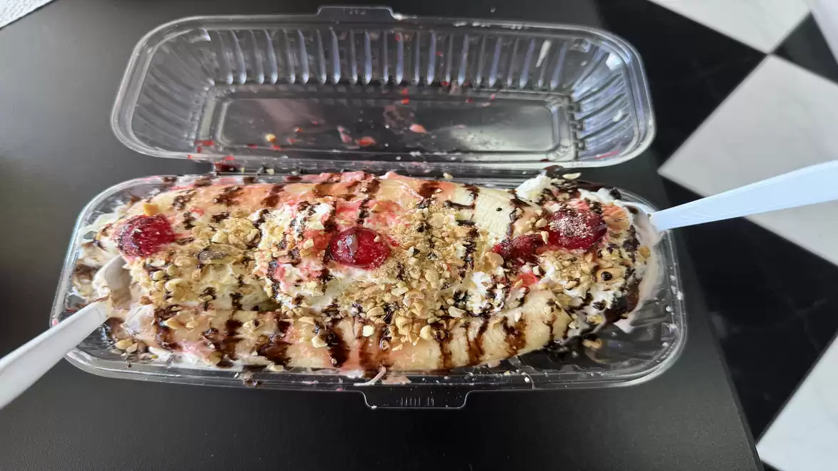 An open plastic container with an ice cream (Bananensplit) specialty garnished with chocolate syrup, nuts, and red fruit pieces. Two plastic spoons are inserted.