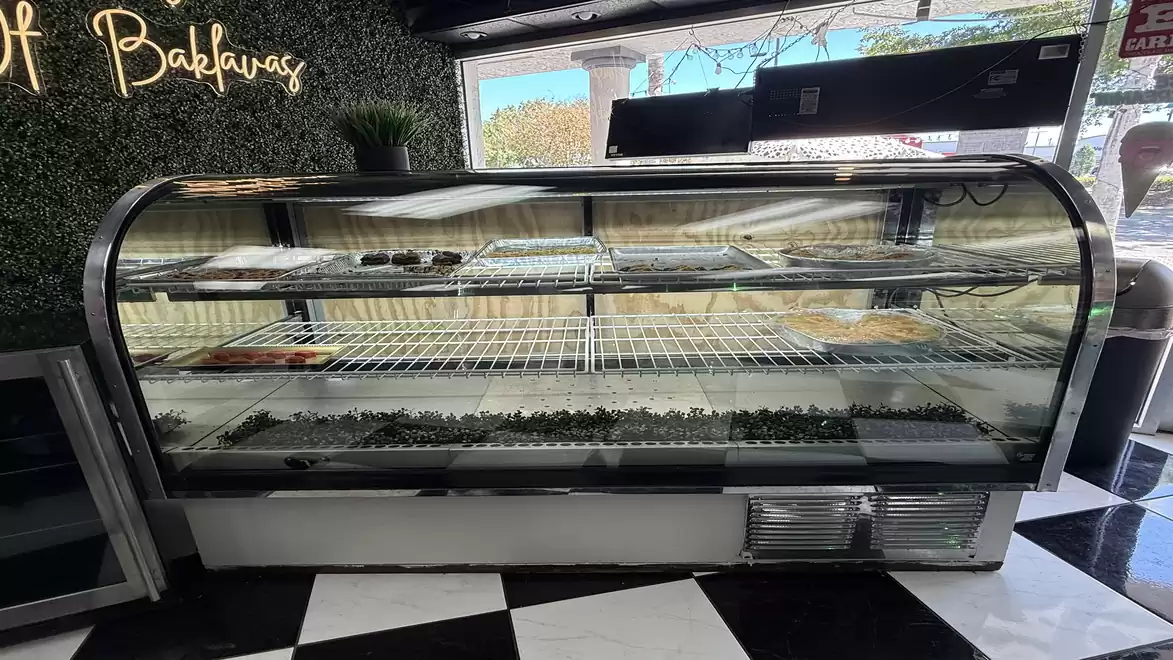 A large, empty display refrigerator in a bakery with 'Baklava' written on the wall, shiny tiles, and sunlight streaming through the window.