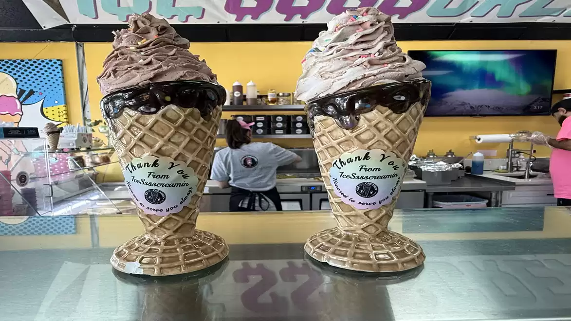 Two large, elaborate ice cream cones filled with soft serve ice cream and chocolate sauce in a colorful ice cream shop.
