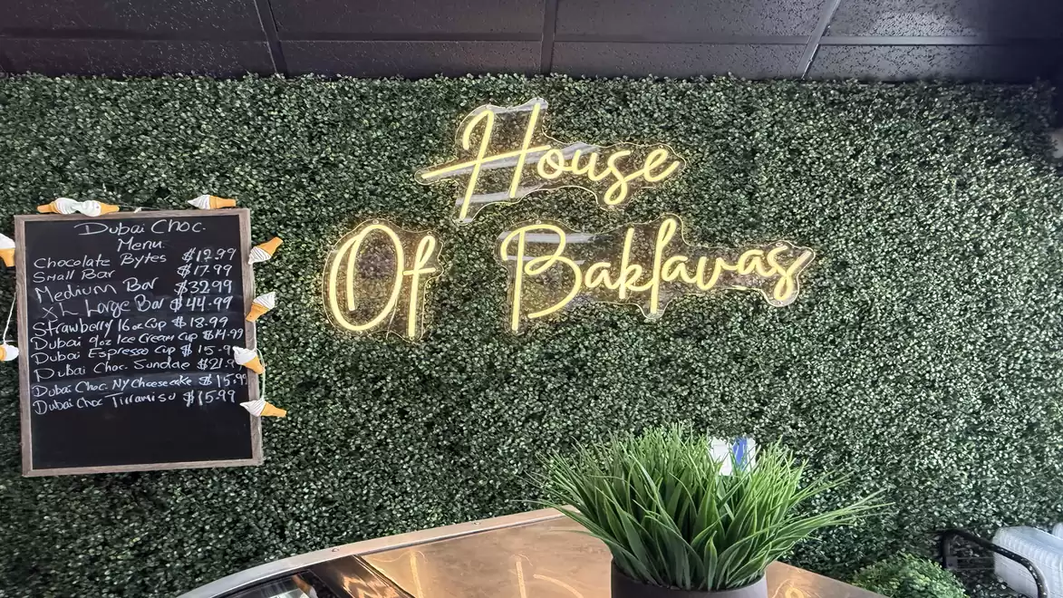 A green background with neon sign 'House Of Baklavas' and a price board with baklava offerings. In the foreground, there is a flower pot.