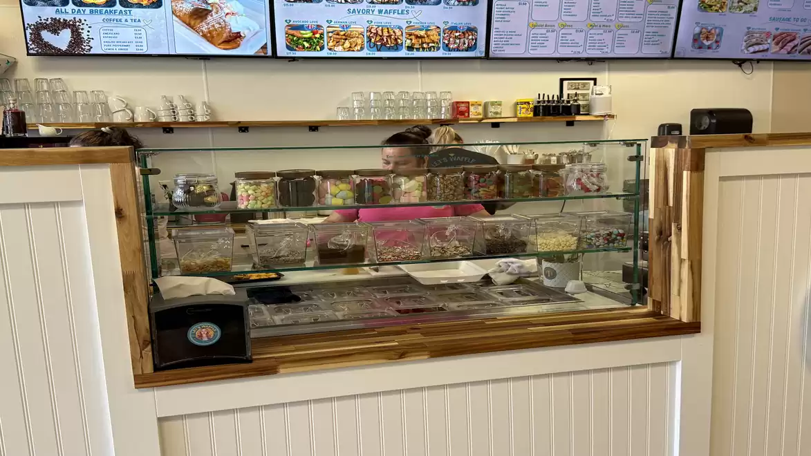 A bakery with a counter, behind which a person is working. The display case contains a variety of sweets and glass containers. In the background, digital menu boards and shelves with jars and products are visible.