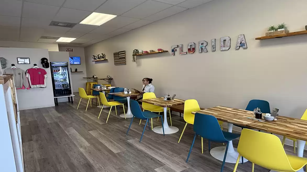 Interior view of a bright café with wooden tables and colorful chairs. The word 'Florida' is displayed on the wall.