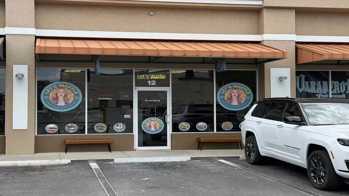 A storefront of Let's Waffle with large windows showcasing the company's logos. A white car is parked to the right of the entrance.