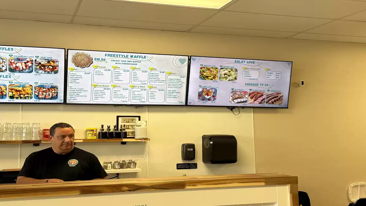 A man behind a counter in a restaurant with digital menus on the wall.