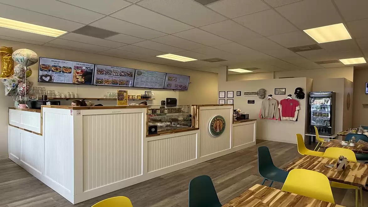 Interior view of a modern waffles restaurant with a white counter, colorful chairs, and wooden tables, illuminated by ceiling lights.
