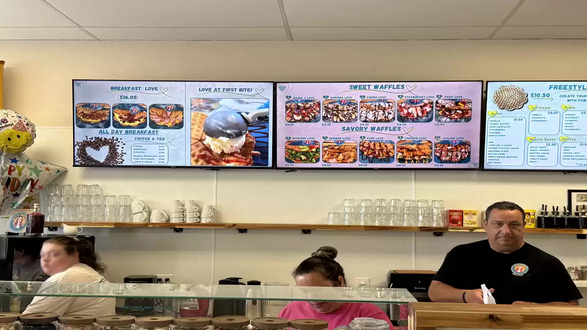 Interior view of a restaurant with three large menu boards on the wall and three people behind the counter. The menu boards display various dishes and prices.