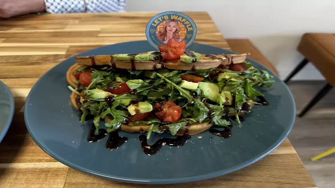 A plate with waffles topped with avocado, arugula, tomatoes, and balsamic glaze, garnished with a small sign.