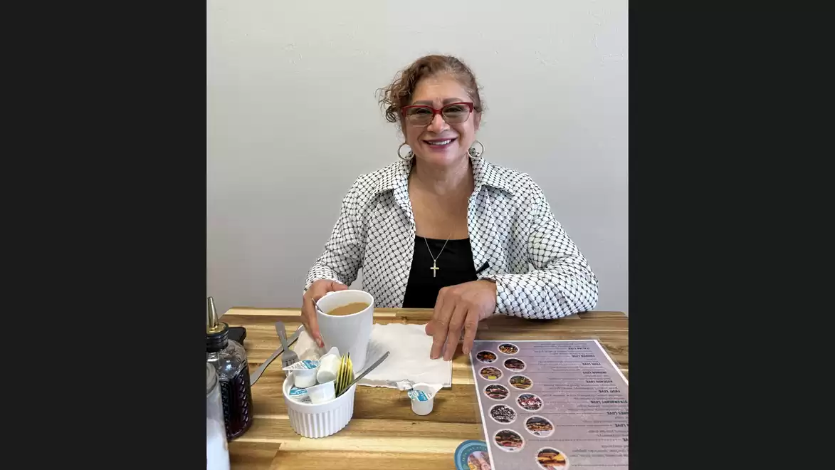 A woman with glasses and a checkered shirt smiles in a café while holding a cup of coffee. A menu and sugar containers are on the table.