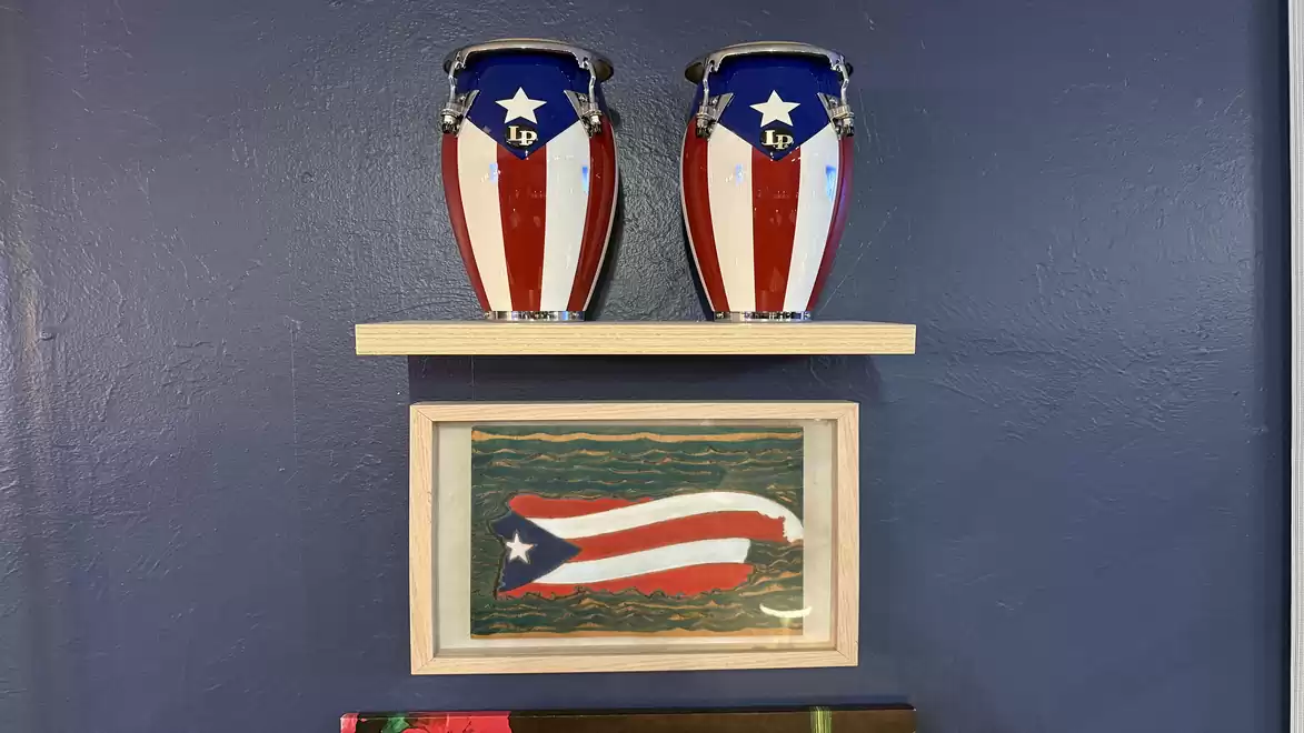 Two congas painted with the Puerto Rican flag stand on a shelf, with a framed picture of the same flag below, all set against a blue wall.