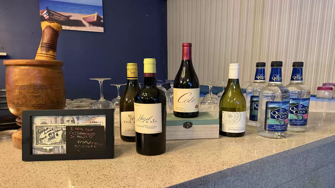 A counter with various wine bottles, vodka bottles, and a wooden mortar, next to a small board with prices.