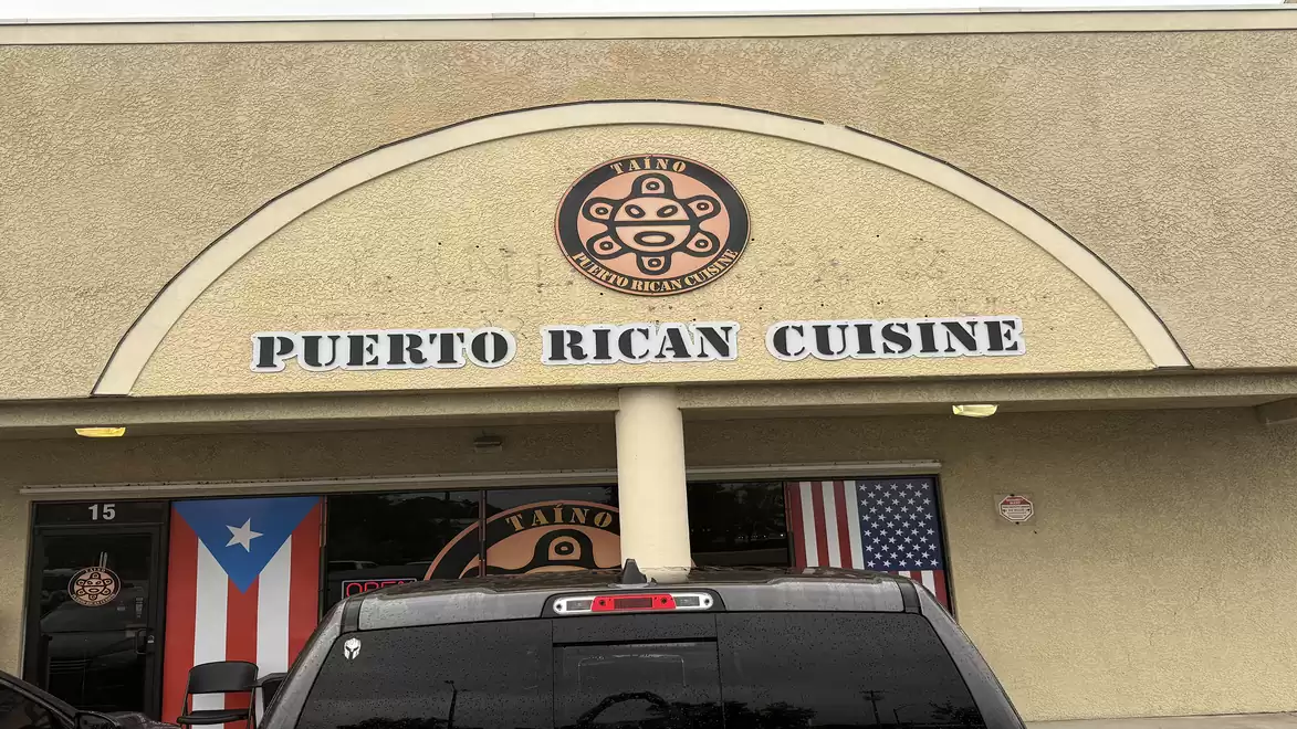Exterior view of a restaurant with the sign 'Puerto Rican Cuisine'. Puerto Rican and American flags flank the entrance.
