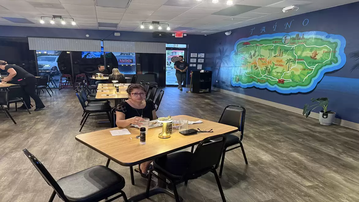 Interior view of a restaurant with tables and chairs where guests are seated. A large mural of Puerto Rico on the background wall. The atmosphere appears welcoming and relaxed.