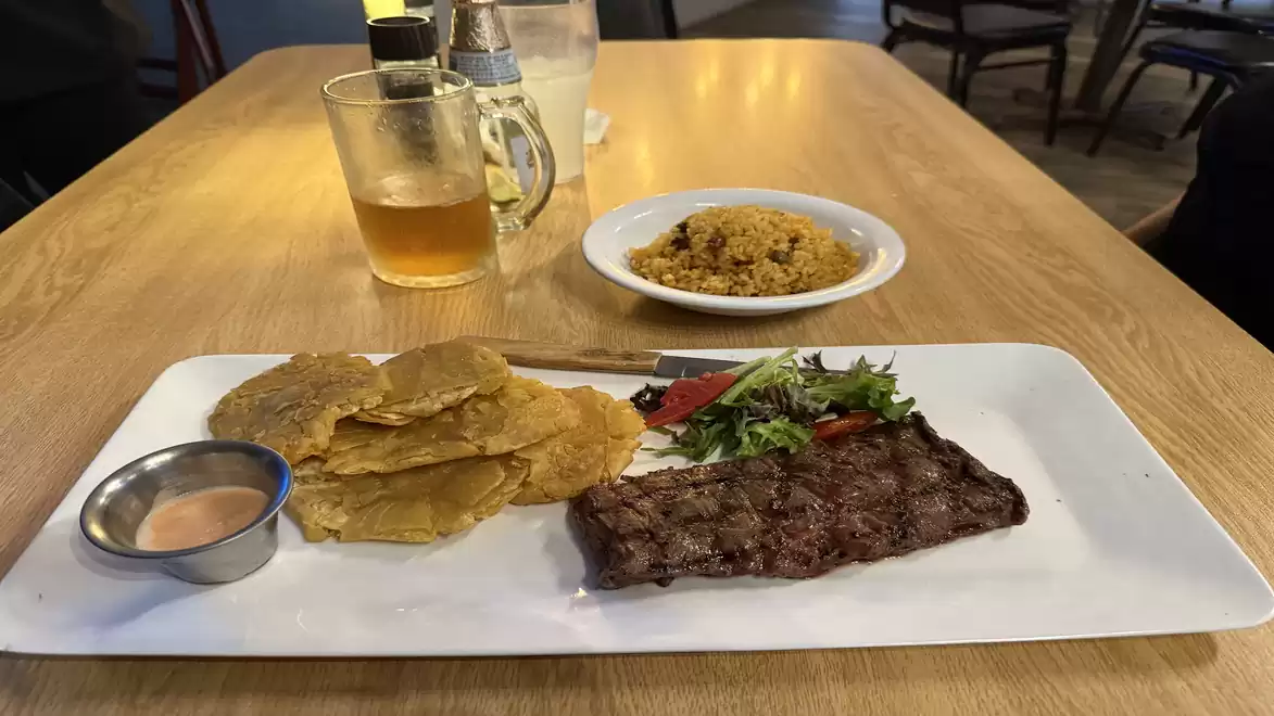 On a wooden table in a restaurant, there is a plate with grilled steak, fried plantain slices, salad, and sauce. Next to it is a glass with a drink and a small plate of rice.
