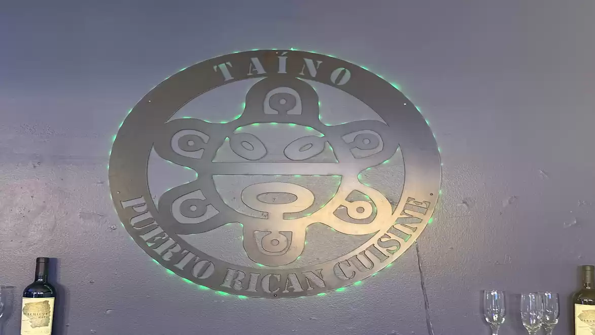 Logo of Taino Puerto Rican Cuisine on a gray wall with wine bottles and glasses underneath.