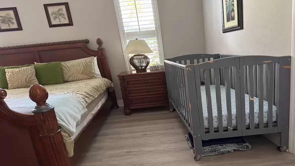 A bedroom with a large wooden bed, a nightstand with lamp, and a gray baby crib on a wooden floor.