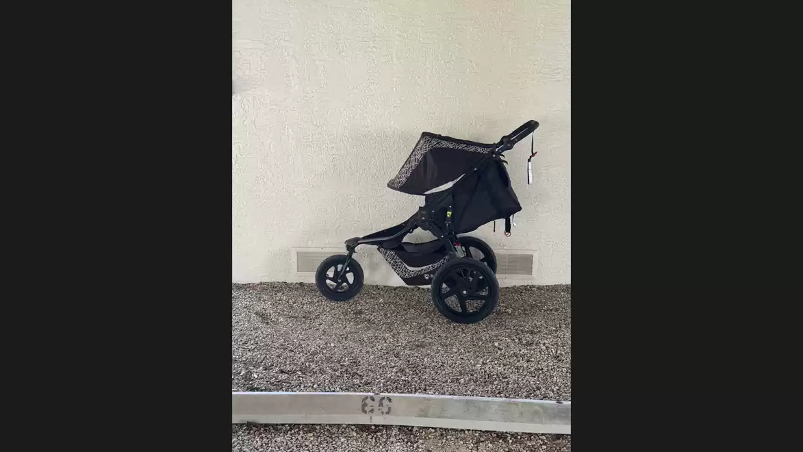 Black stroller parked sideways against a white wall. Gravel ground.