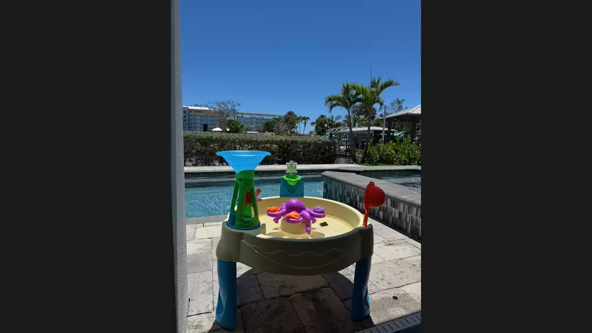 A children's play table with colorful toys, on a patio next to a pool, with palm trees and blue sky in the background.