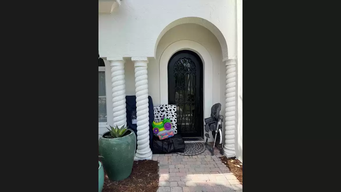 Entrance area of a house with a white, arched door frame and decorative columns. Potted plants and folded garden chairs are in front of the door.