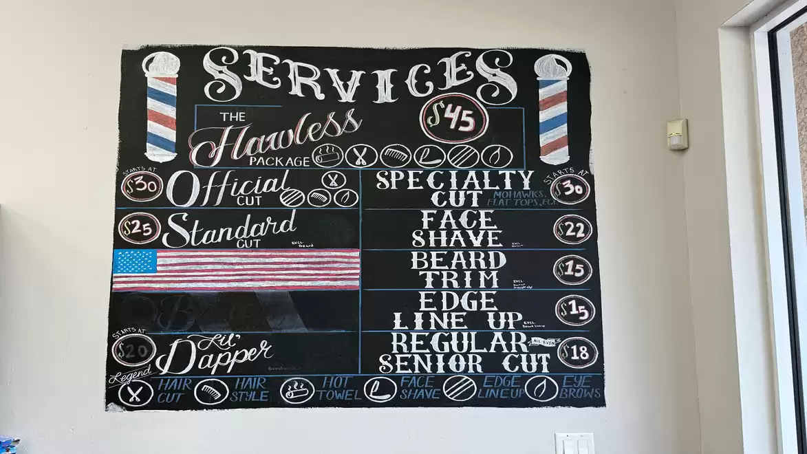 A chalkboard in a barbershop displays prices for various haircut services. The board is artistically designed with handwritten details and decorative elements, including a small drawing of the American flag.