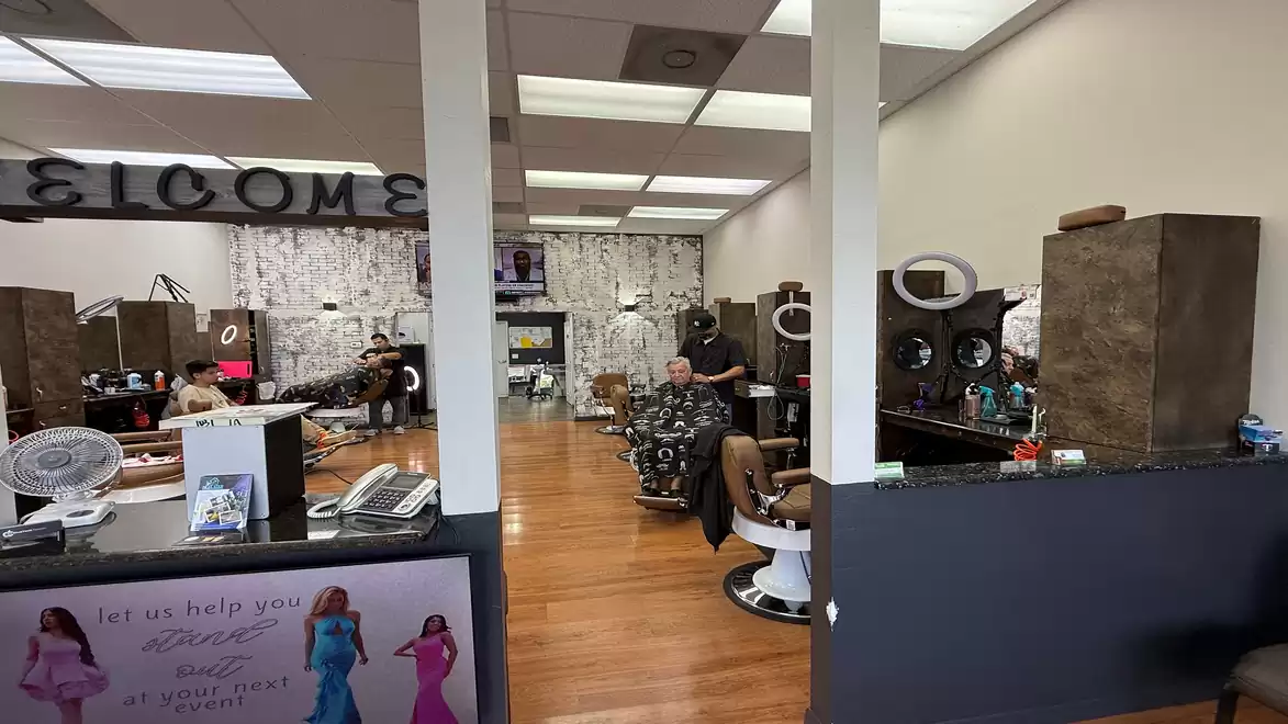 Interior of a hair salon with several barber chairs, customers, and stylists actively working in a busy atmosphere. The decor is modern and welcoming.