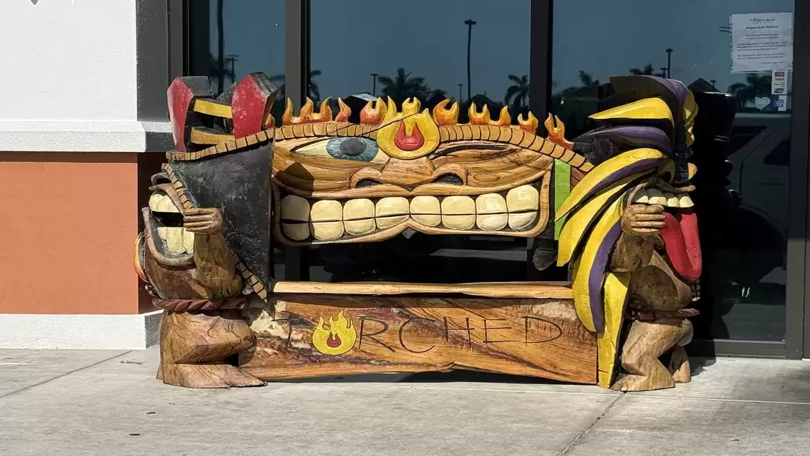 A colorful, artistically carved wooden bench shaped like a smiling face with flame and feather motifs, placed directly in front of a building.
