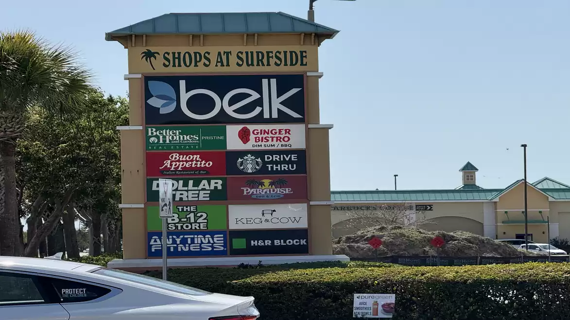 Entrance sign to Shops at Surfside shopping center with several store names on a sunny day.