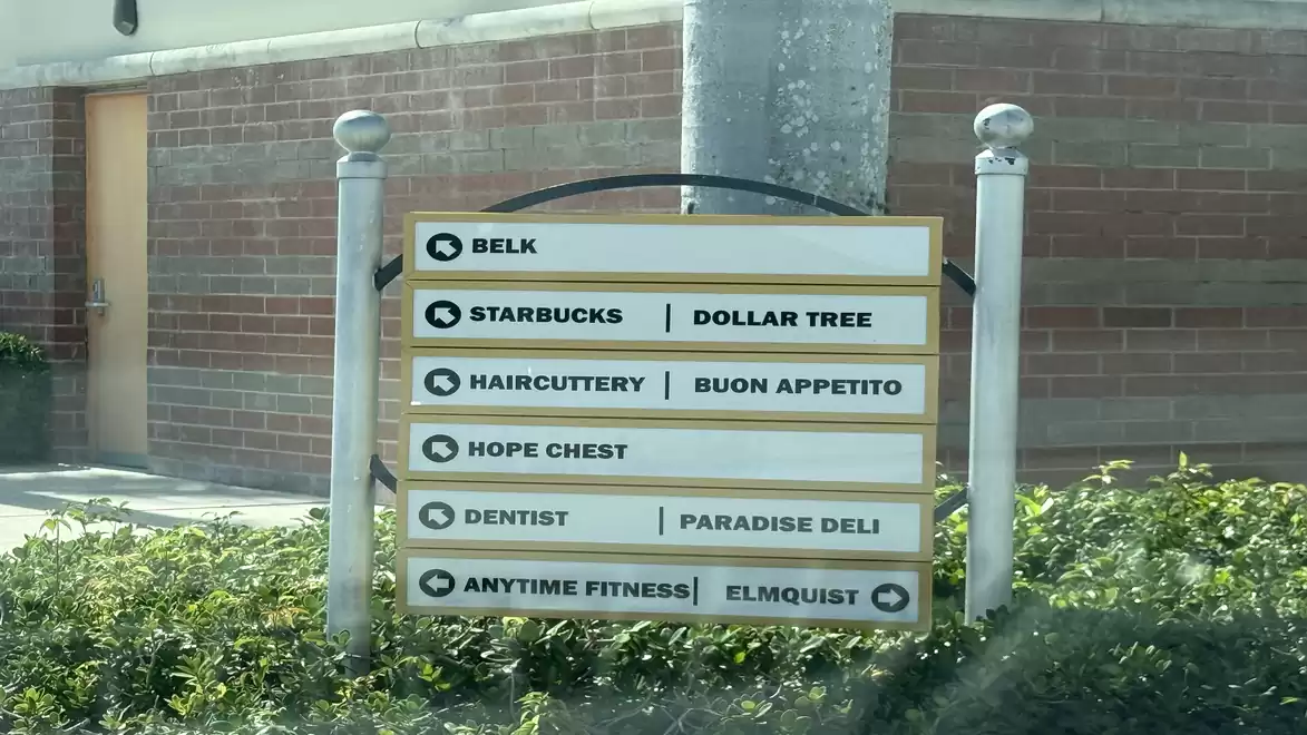An outdoor sign displays the names and directions to various shops and facilities, including Belk, Starbucks, Dollar Tree, Haircuttery, Buon Appetito, Hope Chest, Dentist, Paradise Deli, Anytime Fitness, and Elmquist. The sign is surrounded by plants and 