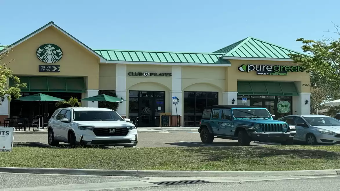 A shopping plaza featuring stores like Starbucks, Club Pilates, and Pure Green. Two cars parked in front of the stores.