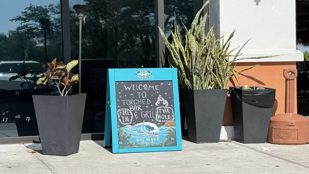 A blue sign in front of a building reading 'Welcome to Torched Bar & Grill' surrounded by potted plants.