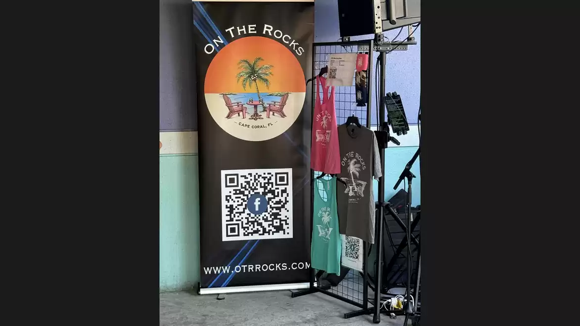 Banner reading 'On The Rocks' with a QR code, next to which T-shirts hang on a grid.